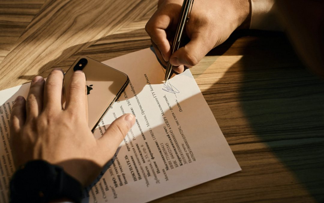 Person holding gold mobile phone in one hand while signing a document with the other